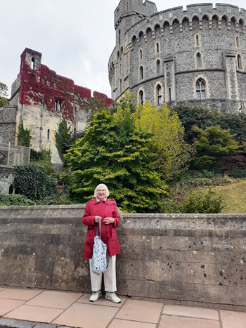 Joan Byrne from Australia standing on a terrace below Windsor Castle's Round Tower. She wears a red coat and holds a patterned bag. Behind her, autumn-coloured trees and ivy-covered walls lead up to the iconic grey stone tower with its crenellated battlements under an overcast sky.