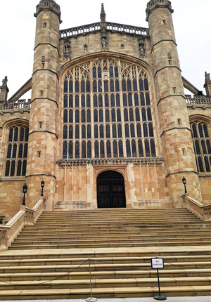 The west front of St George's Chapel at Windsor Castle, viewed from the foot of the entrance steps. The perpendicular Gothic façade features a vast window with intricate stone tracery, flanked by octagonal buttress towers with pinnacles. Stone steps lead up to the arched wooden doorway beneath the window.