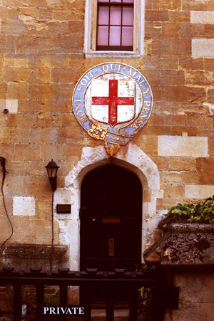 Stone archway entrance to the Military Knights Quarters at Windsor Castle. A heraldic shield displaying the Cross of St George surrounded by the Order of the Garter motto "Honi Soit Qui Mal Y Pense" is mounted above dark wooden double doors. A black iron lantern hangs beside the entrance. A "PRIVATE" sign indicates restricted access.