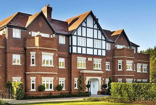 Orchard Lea, a large Edwardian-style residential building in red brick with distinctive black and white Tudor-style half-timbering on the upper gables. The property features multiple bays with white-framed windows, orange clay roof tiles, tall chimneys, and a central arched entrance with a white door. Manicured hedges and topiary shrubs line the front garden.