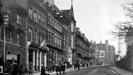 Early 1900s black and white photograph of Sheet Street, Windsor, showing the Royal Albert Institute with its ornate Victorian façade and clock tower. The street scene includes pedestrians, shop fronts with awnings, and period commercial buildings lining both sides. A church or civic building with Gothic features is visible in the distance.