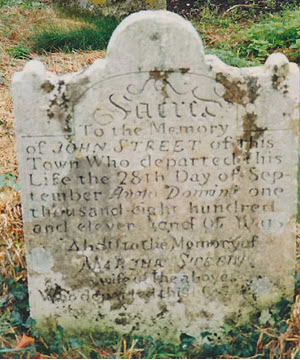 **Alt-text:** Weathered grey stone headstone with decorative curved top and shoulder scrolls. The carved inscription reads "Sacred to the Memory of JOHN STREET of this Town who departed this Life the 28th Day of September Anno Domini one thousand eight hundred and eleven" followed by a memorial to Martha Street, wife of the above. Lichen and moss partially obscure the lower text. Grass surrounds the base.