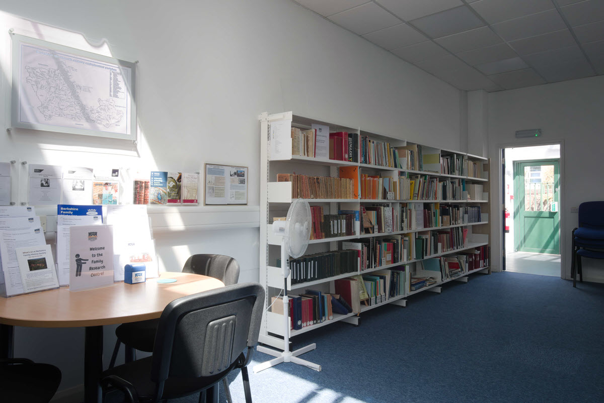 A brightly lit family research library featuring white bookshelves lined with reference books, a round consultation table with informational flyers, and blue carpeted flooring.