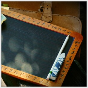 A vintage school slate with the alphabet engraved on its wooden frame, resting with a slate pencil in front of a leather satchel.
