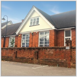 A red brick building with a prominent cream-colored gable and large multi-pane windows, situated behind a low brick boundary wall.