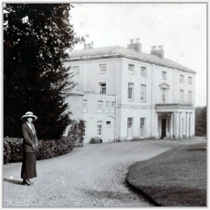 A woman in early 20th-century attire stands on a gravel driveway in front of a large, three-story Georgian-style manor house with a columned portico.