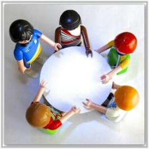 Five diverse toy figures stand in a circle around a blank white round table, reaching their hands towards the center.