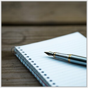 A silver fountain pen with gold accents rests on an open, lined spiral notebook on a wooden surface.