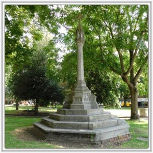 A historic stone cross monument with a tiered octagonal base, situated in a grassy park surrounded by lush green trees.