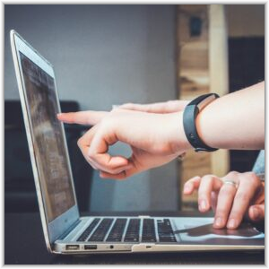 Two people collaborating on a laptop, with one person pointing at the screen and another using the trackpad.