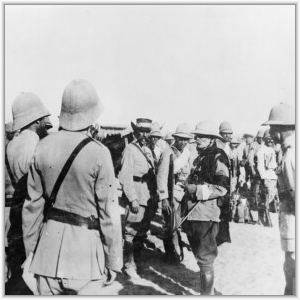 A black and white historical photograph of military officers in khaki uniforms and pith helmets conversing in a desert setting.