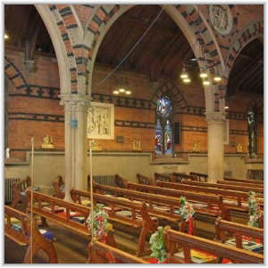 Interior view of a church showing wooden pews decorated with floral arrangements, stone pillars supporting brick arches, and stained-glass windows in the background.