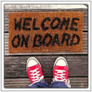 Overhead view of a person wearing red canvas sneakers standing on wooden decking in front of a doormat that reads "WELCOME ON BOARD.