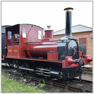 A vintage maroon and black steam locomotive, marked with yellow lettering as No. 5, sitting on railway tracks next to a brick building.