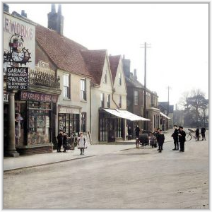 A historical street scene showing village shopfronts, including Charles G. Brown's store and a cycle works, with children in early 20th-century attire standing along the road.